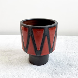 Könnte beinhalten: Eine Keramikvase mit geometrischem Design. Die Vase hat ein rot-schwarzes Zickzackmuster um den Körper und einen soliden schwarzen Sockel. Der Innenraum ist dunkelbraun. Die Vase ist ein dekorativer Gegenstand.