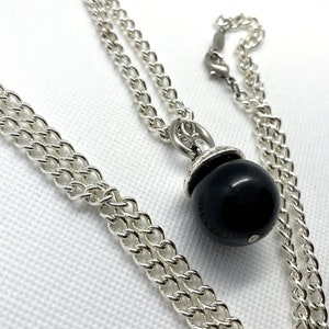 Op de afbeelding: Een zilveren ketting met een zwarte, bolvormige hanger. De ketting is een dubbele streng van zilveren schakels. De hanger is een glanzende zwarte bol met een zilveren dop. De ketting wordt op een witte ondergrond gepresenteerd.