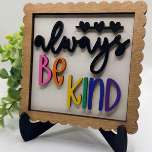 以下が含まれることがあります： 「always be kind」の文字が黒、ピンク、オレンジ、黄色、緑、紫で書かれた木製のサイン。サインはスカラップエッジで、黒いスタンドで支えられています。「always」という単語の上には3つの黒いハートがあります。