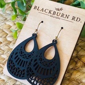 May include: Blackburn Rd. Designs earrings are black wooden teardrop shaped with a delicate laser cut design. The earrings are hanging on a wooden display card.