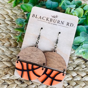 Peut inclure: Une paire de boucles d'oreilles en bois avec un motif de ballon de basket. Les boucles d'oreilles sont orange et noires avec une finition en bois. Les boucles d'oreilles sont sur une carte en bois avec le texte "Blackburn Rd. Designs".