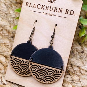 Peut inclure: Une paire de boucles d'oreilles en bois avec une finition noire et bois naturel. Les boucles d'oreilles présentent un motif géométrique de vagues qui se chevauchent. Les boucles d'oreilles sont présentées sur une carte en bois avec le texte "Blackburn Rd. Designs".