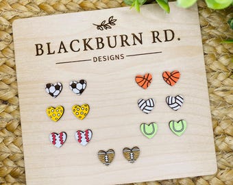 以下が含まれることがあります： 「Blackburn Rd. Designs」という文字が書かれた木製のディスプレイボードには、さまざまなスポーツをテーマにしたハート型のイヤリングが展示されています。イヤリングには、バスケットボール、サッカーボール、バレーボール、野球、アメリカンフットボール、テニスラケットのデザインが施されています。