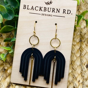 May include: Black and gold geometric earrings with a modern design. The earrings feature a series of black arches with a gold bar hanging from the bottom. The earrings are on a wooden display card with the text "Blackburn Rd. Designs".