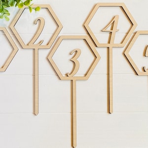 Può includere: Cinque porta numeri da tavolo esagonali in legno con finitura in legno naturale. Ogni porta numeri ha un numero da 1 a 5 ritagliato al centro. I numeri sono scritti in un carattere corsivo.