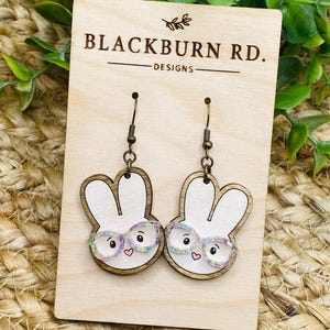May include: A pair of wooden bunny rabbit earrings with iridescent glitter glasses. The earrings are on a wooden display card with the text "Blackburn Rd. Designs".