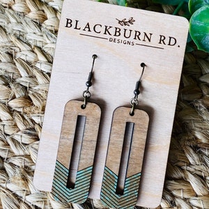 May include: A pair of wooden earrings with a teal and brown geometric design. The earrings are on a wooden display card with the text "Blackburn Rd. Designs".