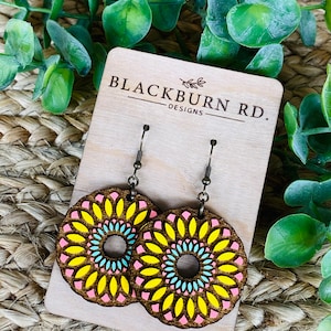 May include: A pair of wooden earrings with a pink, yellow, and blue mandala design. The earrings are on a wooden display card with the text "Blackburn Rd. Designs".