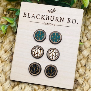 May include: A set of six wooden earrings with different patterns. The earrings are round and have a black, white, and teal color scheme. The earrings are displayed on a wooden card with the text "Blackburn Rd. Designs".
