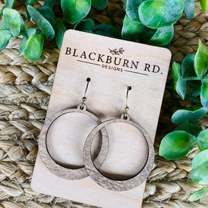 Op de afbeelding: Een paar houten creolen met een lichtbruine houtnerf. De oorbellen worden gepresenteerd op een houten kaart met de tekst "Blackburn Rd. Designs".