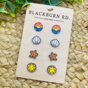 May include: A set of six wooden earrings with summer-themed designs. The earrings feature a rainbow sunset, a seashell, a starfish, and a smiling sun. The earrings are displayed on a wooden card with the text "Blackburn Rd. Designs".