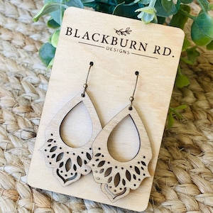 May include: A pair of wooden earrings with a delicate, intricate design. The earrings are light brown in color and have a teardrop shape. The earrings are displayed on a wooden card with the text "Blackburn Rd. Designs".