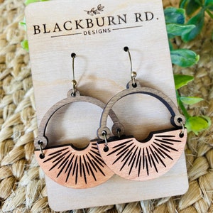 May include: A pair of wooden earrings with a sunburst design. The earrings are brown and have a gold-tone hook. The earrings are displayed on a wooden card with the text "Blackburn Rd. Designs".