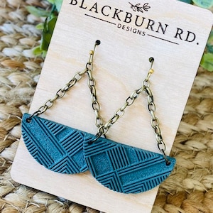 May include: Teal blue geometric patterned half-circle earrings with gold chain links and brass hooks. The earrings are displayed on a wooden card with the text "Blackburn Rd. Designs".