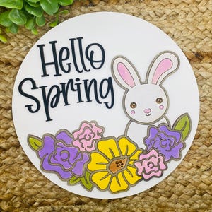 Puede incluir: Un letrero redondo de madera blanca con el texto "Hello Spring" en letras negras. Un conejito blanco con orejas rosas asoma por detrás de un grupo de flores moradas, rosas y amarillas.
