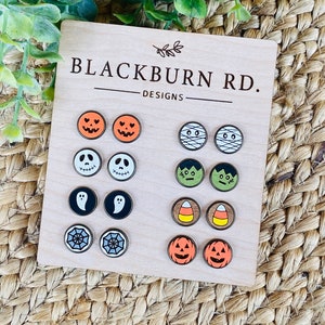 May include: A set of 10 Halloween-themed wooden earrings. The earrings feature various designs, including pumpkins, ghosts, mummies, spiders, and candy corn. The earrings are displayed on a wooden board with the text "Blackburn Rd. Designs".