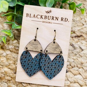 May include: A pair of wooden earrings with a geometric heart design. The earrings are blue with a white pattern and have a wood grain finish. The earrings are on a wooden display card with the text "Blackburn Rd. Designs".