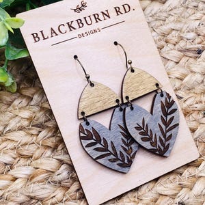 Puede incluir: Un par de pendientes de madera con un diseño de corazón gris con un patrón de hojas. Los pendientes están unidos a ganchos de color dorado y se muestran en una tarjeta de madera con el texto "Blackburn Rd. Designs".