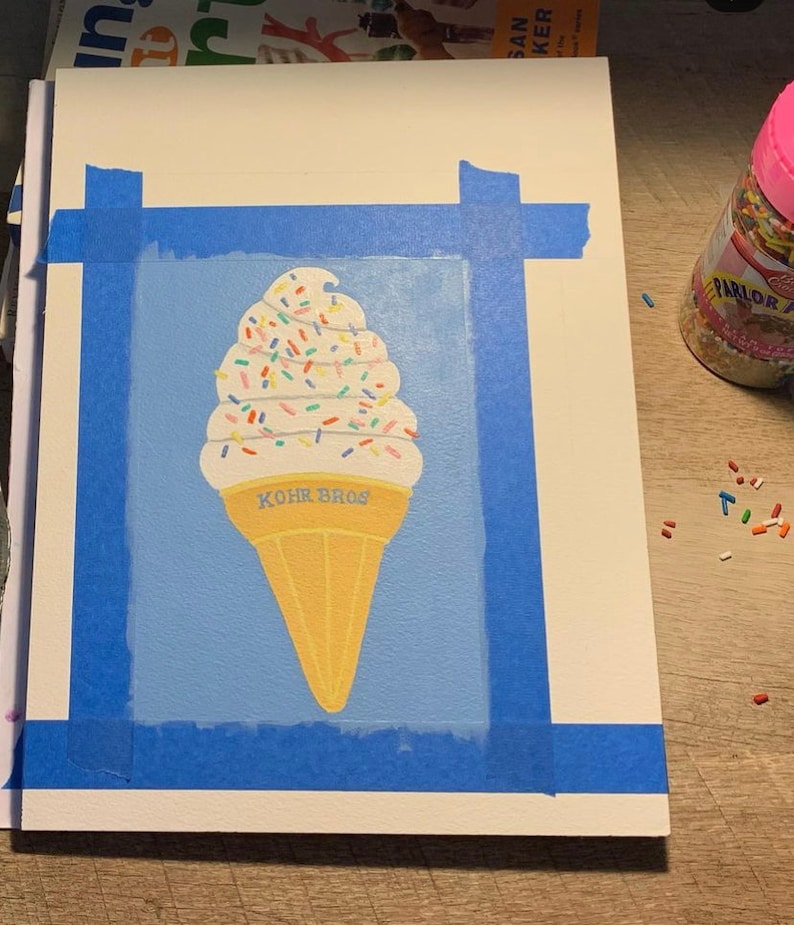 Pu&ograve; includere: Dipinto di un cono gelato su sfondo azzurro, fissato a una superficie bianca con nastro adesivo blu. Il gelato ha un gelato soft bianco con granelli colorati e un cono giallo. Il testo "KOHR BROS" &egrave; sul cono.