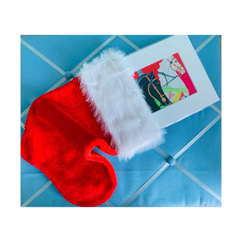 K&ouml;nnte beinhalten: Ein roter Weihnachtsstrumpf mit wei&szlig;em Kunstfellbesatz, neben einem gerahmten Druck von zwei Karussellpferden. Der Druck zeigt ein schwarzes und ein wei&szlig;es Pferd mit farbenfrohen Akzenten. Die Artikel befinden sich auf einer hellblauen Oberfl&auml;che.