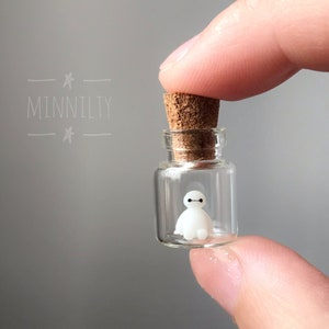 Può includere: Un piccolo barattolo di vetro con un tappo di sughero contiene una statuetta bianca di Baymax, il robot gonfiabile del film Disney Big Hero 6.