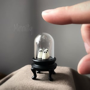 以下が含まれることがあります： 黒いガラスのクロッシュの中に、白い幽霊の置物が3つ入っています。クロッシュは、3本の脚が付いた黒い台座の上に置かれています。クロッシュは、茶色の表面の上に置かれています。