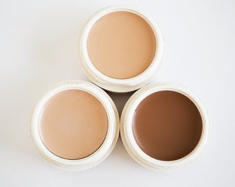 May include: Three round containers of concealer, each with a different shade of foundation. The shades are light, medium, and dark.