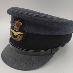 Könnte beinhalten: Ein dunkelblauer Hut im Militärstil mit einem schwarzen Lederband und einem goldenen gestickten Emblem mit einer Krone und einem geflügelten Adler.