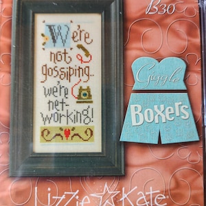 May include: A framed cross-stitch sampler with the text "We're not gossiping, we're networking!" in various colors. A light blue cutout of a pair of boxers with the word "Boxers" is next to the frame. The brand name "Lizzie Kate" is at the bottom.