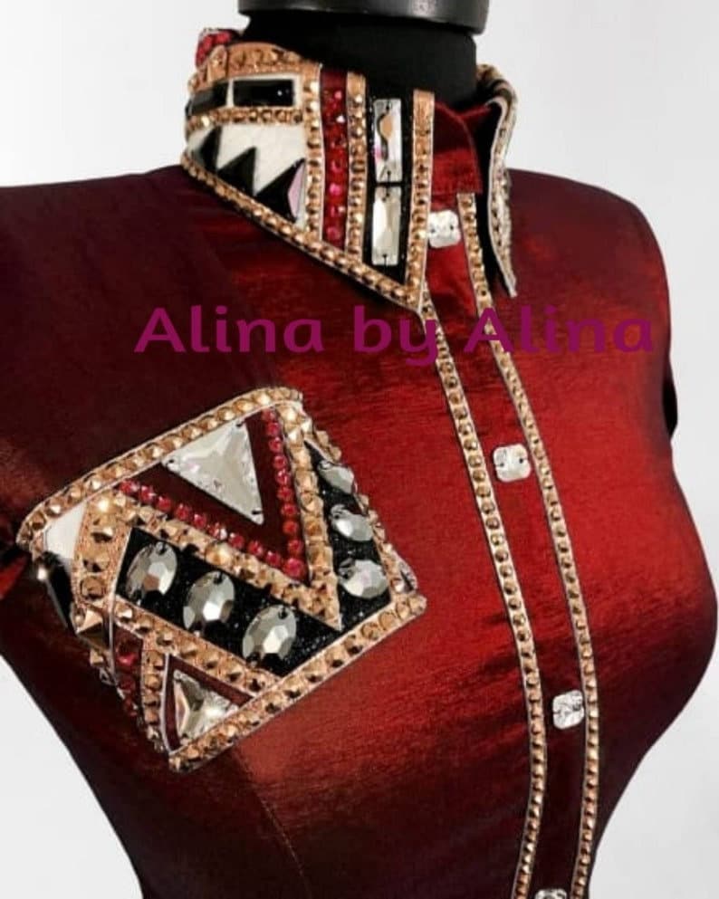 K&ouml;nnte beinhalten: Ein burgunderfarbenes Reitshirt mit einem gold- und schwarzbesetzten Kragen und einer passenden, besetzten &Auml;rmelmanschette. Das Shirt tr&auml;gt den Text "Alina by Alina" auf der Vorderseite.