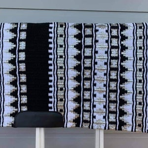 May include: A rectangular horse saddle pad with a black, white, and silver geometric pattern. The pad has a black spine and a black leather-like piece at the bottom. The pad is displayed on a white stand.