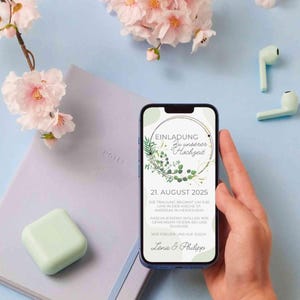 May include: A smartphone displaying a wedding invitation with a green and white floral design. The invitation reads "Einladung zu unserer Hochzeit" and lists the date "21. August 2025".