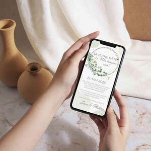 May include: A smartphone displaying a save the date announcement for a wedding. The announcement features a green wreath with the text "Save the Date" and "We're getting married!" The date is May 21, 2025, and the names Lena and Philipp are at the bottom.