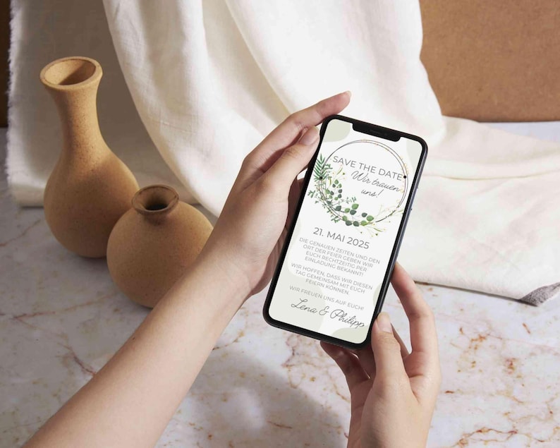 May include: A smartphone displaying a save the date announcement for a wedding. The announcement features a green wreath with the text "Save the Date" and "We're getting married!" The date is May 21, 2025, and the names Lena and Philipp are at the bottom.