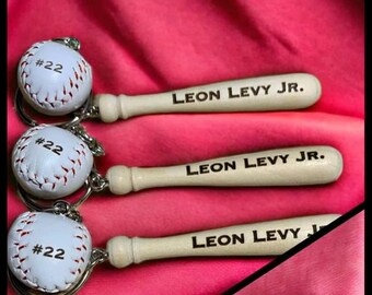 May include: Baseball-themed keyrings featuring white baseballs and wooden bat handles. The baseballs have the number "#22" or "#12" printed on them. The bat handles are engraved with the names "LEON LEVY JR." or "KENNEDY".