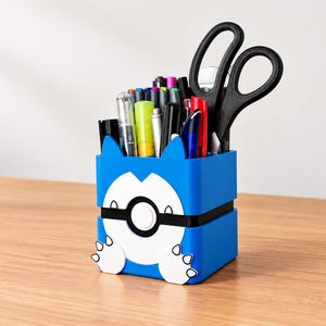 Könnte beinhalten: Ein blauer, quadratischer Schreibtisch-Organizer mit einem weiß-schwarzen, kreisförmigen Emblem auf der Vorderseite, das an einen Ball erinnert, sowie zwei kleinen, weißen, krallenartigen Akzenten an der Basis. Der Behälter ist mit verschiedenen Büroartikeln gefüllt.
