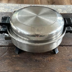 Peut inclure: Une poêle électrique vintage avec une surface de cuisson en acier inoxydable et un couvercle en forme de dôme. La poêle a des poignées noires et repose sur des pieds noirs. L'appareil est conçu pour la cuisine et est placé sur une surface en bois.