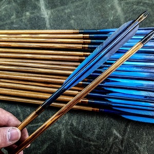 May include: A set of wooden arrows with blue and black fletching. The arrows are arranged in a stack, with the fletching facing up. The arrows are held in a hand.