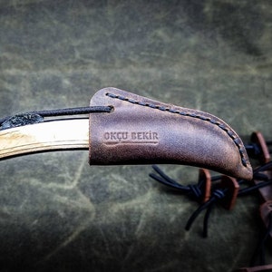 May include: A close-up shot of a traditional recurve bow, featuring a brown leather bow tip protector. The protector is embossed with the text "OKÇU BEKIR". Black cords and leather details are visible. The bow has a natural wood grain finish.