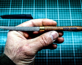 May include: A metal rasp tool with a textured, pointed blade and a polished brass ferrule connecting to a dark brown wooden handle. The handle features the engraved text "BACON BAKER" in a bold, sans-serif font. A black protective sheath is visible.