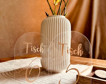 May include: Two clear acrylic table number holders with gold lettering. The holders are shaped like arches and have the words "Tisch" and the numbers "6" and "5" written on them. The holders are sitting on a table with a white tablecloth and a vase of yellow flowers.
