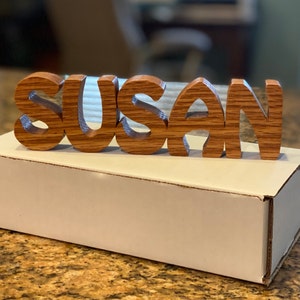 May include: Wooden name sign spelling "SUSAN" in a decorative font. The letters are made of light brown wood with a grain pattern. The sign is sitting on a white box.
