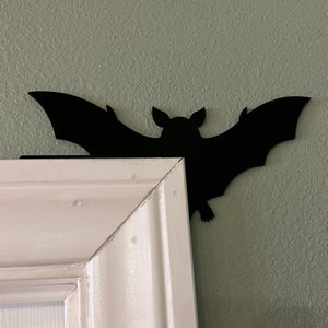 Puede incluir: Decoraci&oacute;n de pared con silueta de murci&eacute;lago negro, montada en un marco de puerta blanco. El murci&eacute;lago est&aacute; hecho de metal y tiene un dise&ntilde;o simple y elegante.