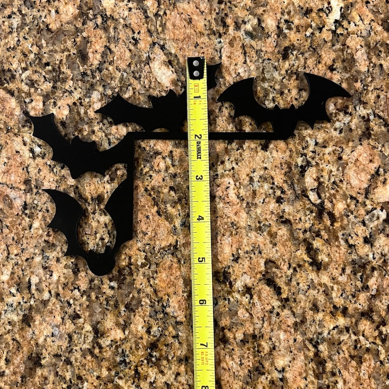 Puede incluir: Decoraci&oacute;n de pared de metal negro en forma de silueta de murci&eacute;lago con cuatro murci&eacute;lagos dispuestos en un racimo. La decoraci&oacute;n se mide con una cinta m&eacute;trica amarilla que indica 20 cm.