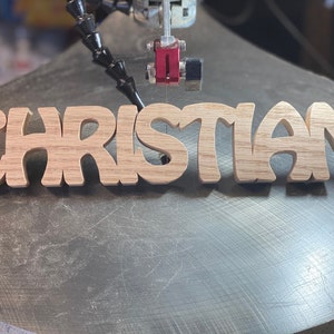 May include: A close-up of a wooden sign being cut out on a scroll saw. The sign reads "CHRISTIAN" in a stylized font.