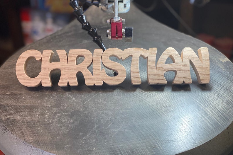 May include: A close-up of a wooden sign being cut out on a scroll saw. The sign reads "CHRISTIAN" in a stylized font.
