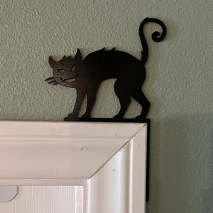 Puede incluir: Silueta de gato de metal negro con cola enroscada, montada en un marco de puerta blanco.