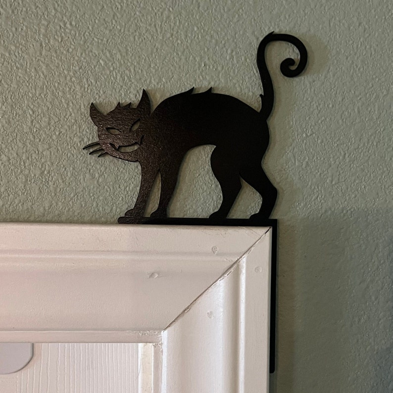 Puede incluir: Silueta de gato de metal negro con cola enroscada, montada en un marco de puerta blanco.