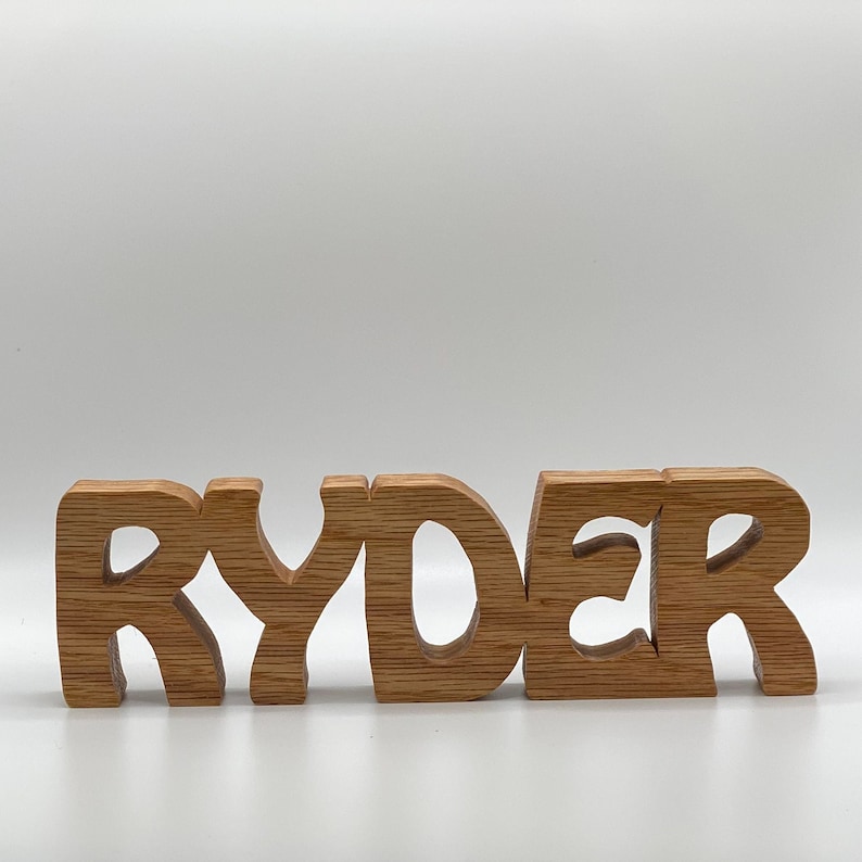 May include: Wooden name sign spelling "RYDER" in a whimsical font. Each letter is individually carved and stands on its own. The sign is made of light brown wood.