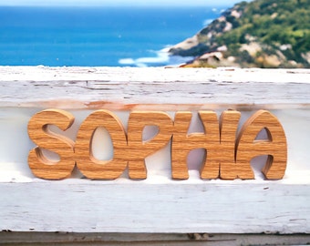 May include: Wooden letters spelling out the name "Sophia" in a light brown color. The letters are arranged in a row on a white wooden surface. The background is a blurry image of a blue ocean and green hills.